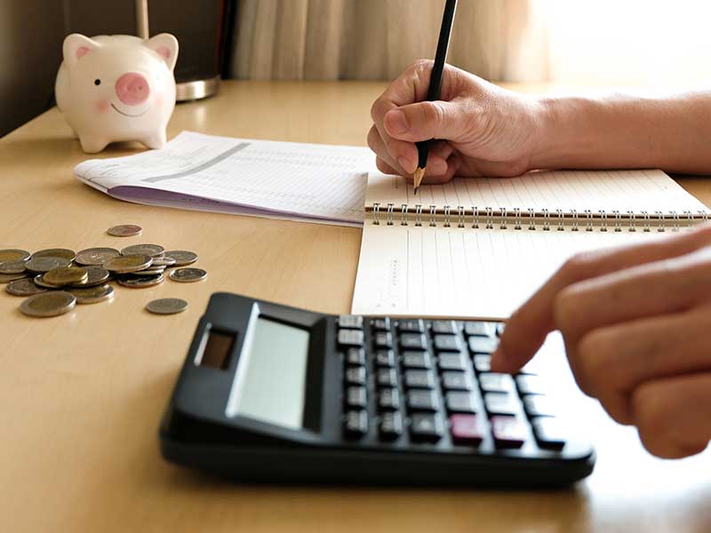 Close up of a desk with a piggybank and a person's hands using a calculator to do some financing while writing it down